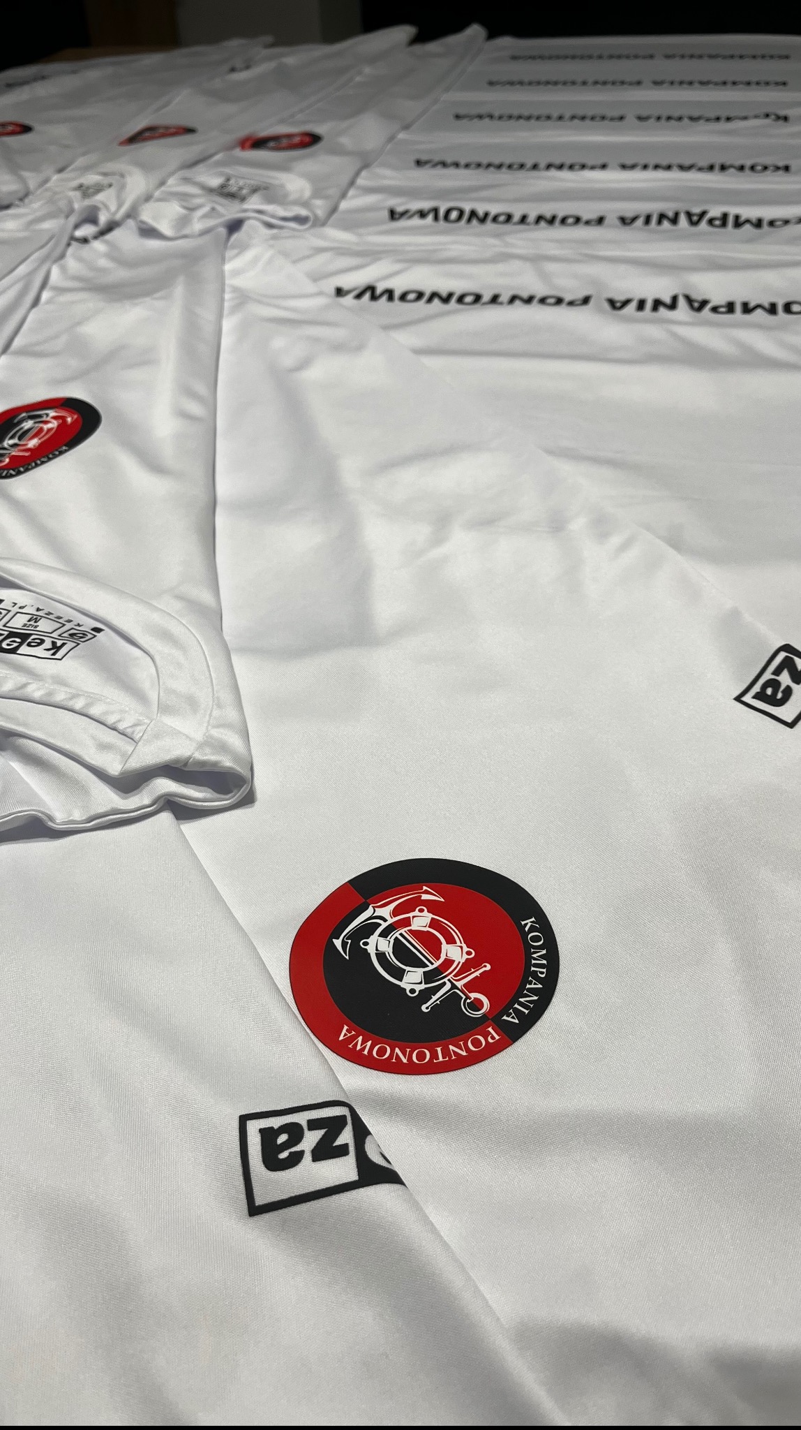 Białe koszulki z nadrukiem logo 'Kompania Pontonowa' i małym logo 'za' w rogu, ułożone warstwami, widoczne detale nadruku i materiału.