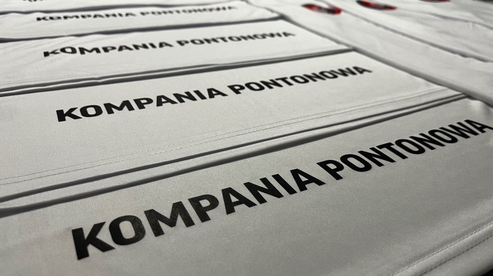 Białe koszulki z czarnym nadrukiem 'KOMPANIA PONTONOWA' ułożone rzędami, widoczne szwy i faktura materiału. Perspektywa z góry, ostre światło.