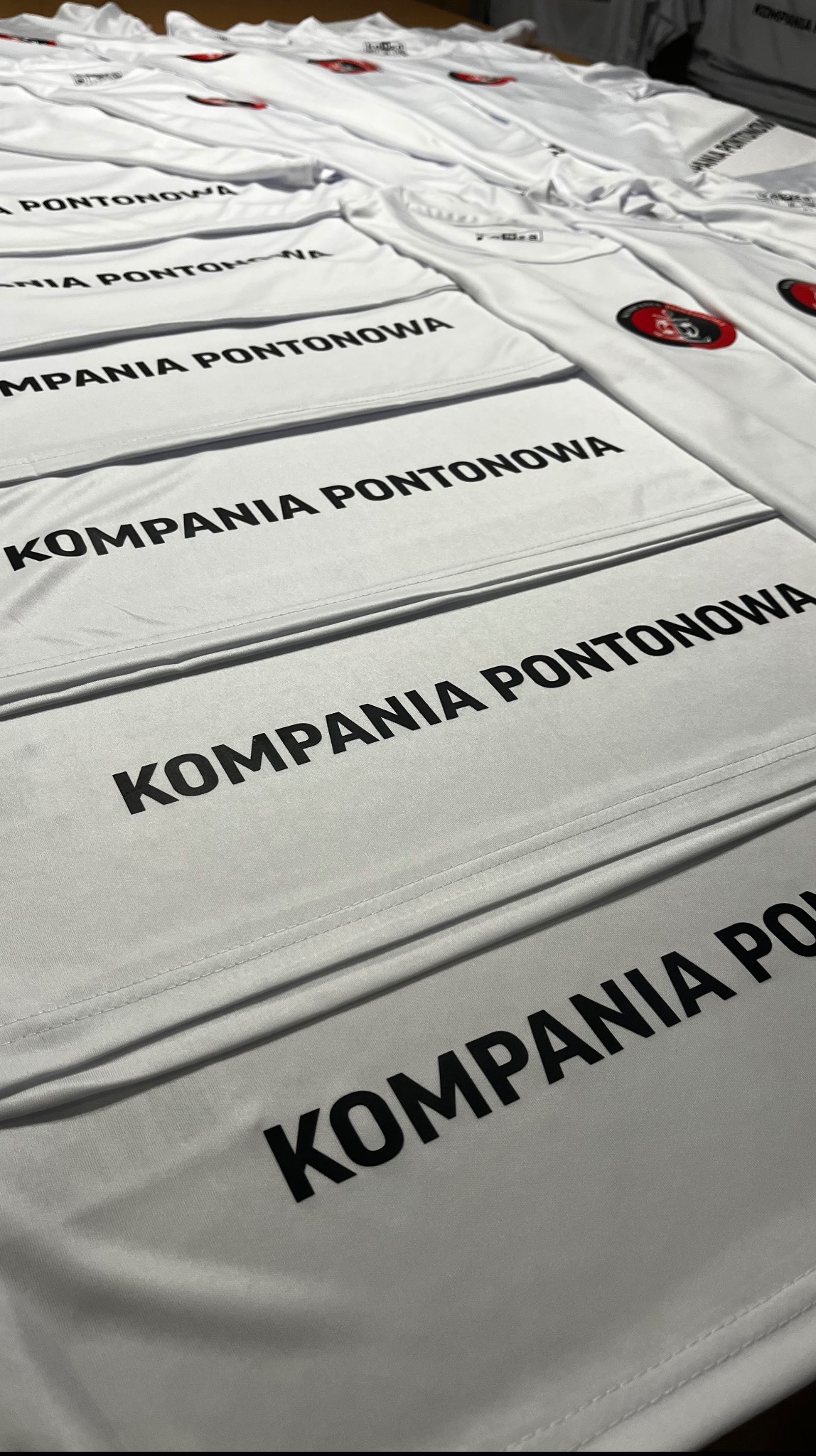Stos białych koszulek z czarnym nadrukiem 'KOMPANIA PONTONOWA' i logo. Ułożone równo, widoczne szwy i detale materiału. Perspektywa z góry, oświetlenie studyjne.