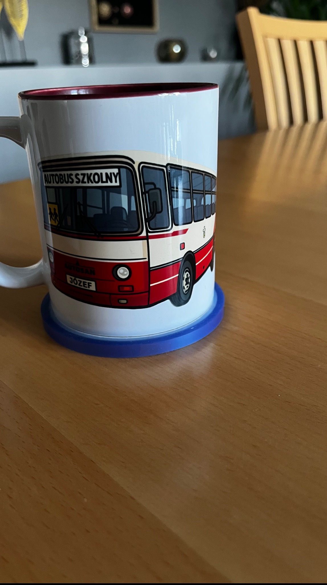 Ceramiczny kubek z nadrukiem autobusu szkolnego, stojący na niebieskiej podkładce na drewnianym stole. Kubek ma czerwone obramowanie i napis 'Autobus Szkolny' oraz imię 'Józef'.