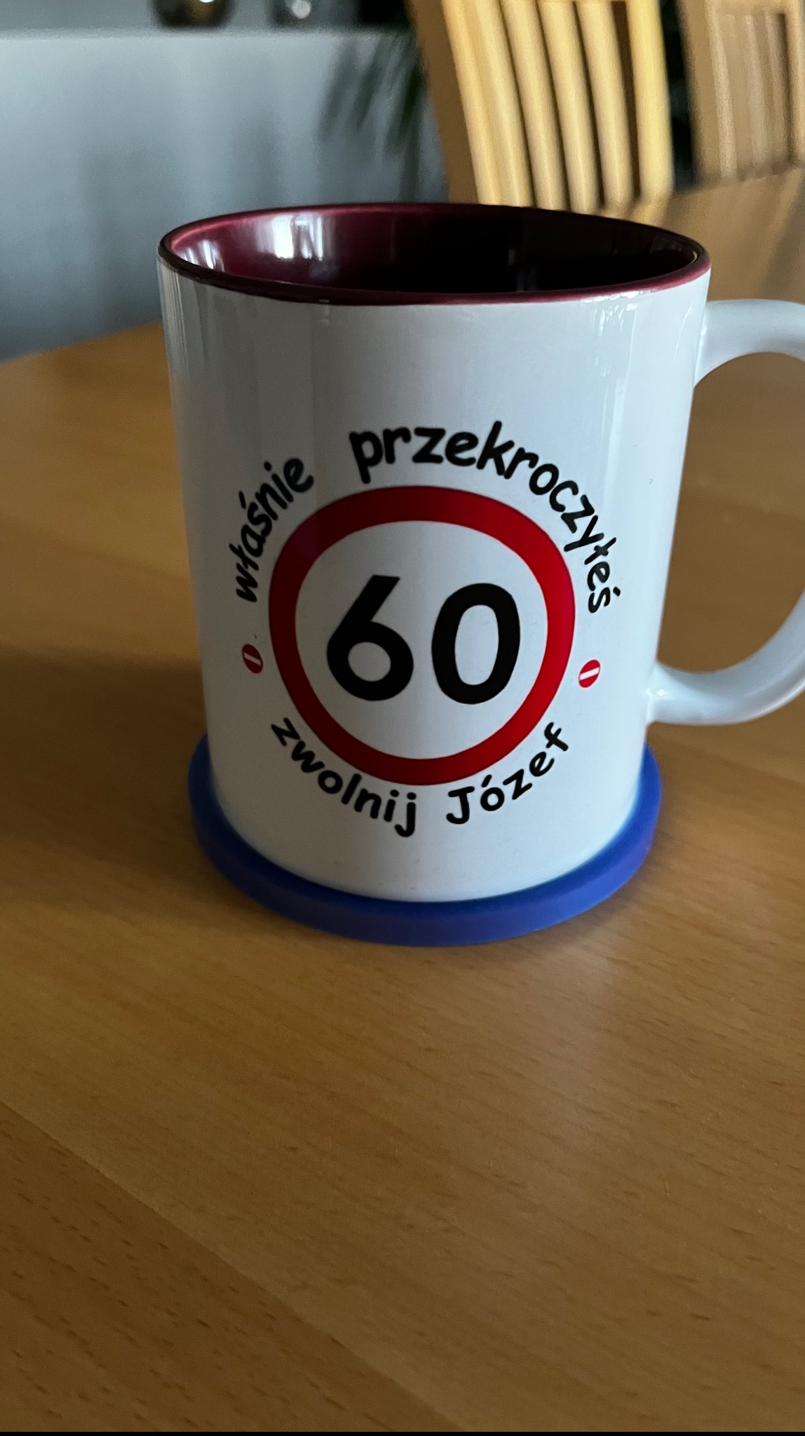 Biały kubek z czerwonym wnętrzem, na niebieskiej podkładce. Na kubku nadruk: 'właśnie przekroczyłeś 60 zwolnij Józef' w formie znaku drogowego.