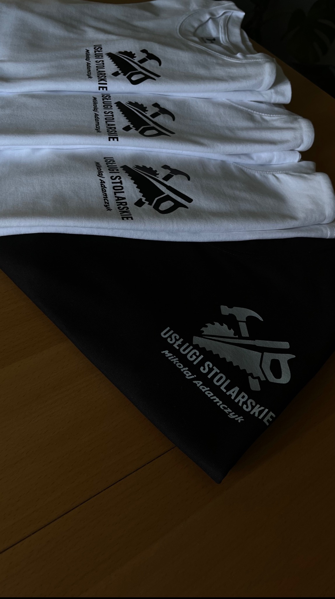 Stos białych koszulek i czarna torba z nadrukiem logo 'Usługi Stolarskie Mikołaj Adamczyk', prezentacja gotowych produktów z branży odzieżowej z unikalnym wzorem.