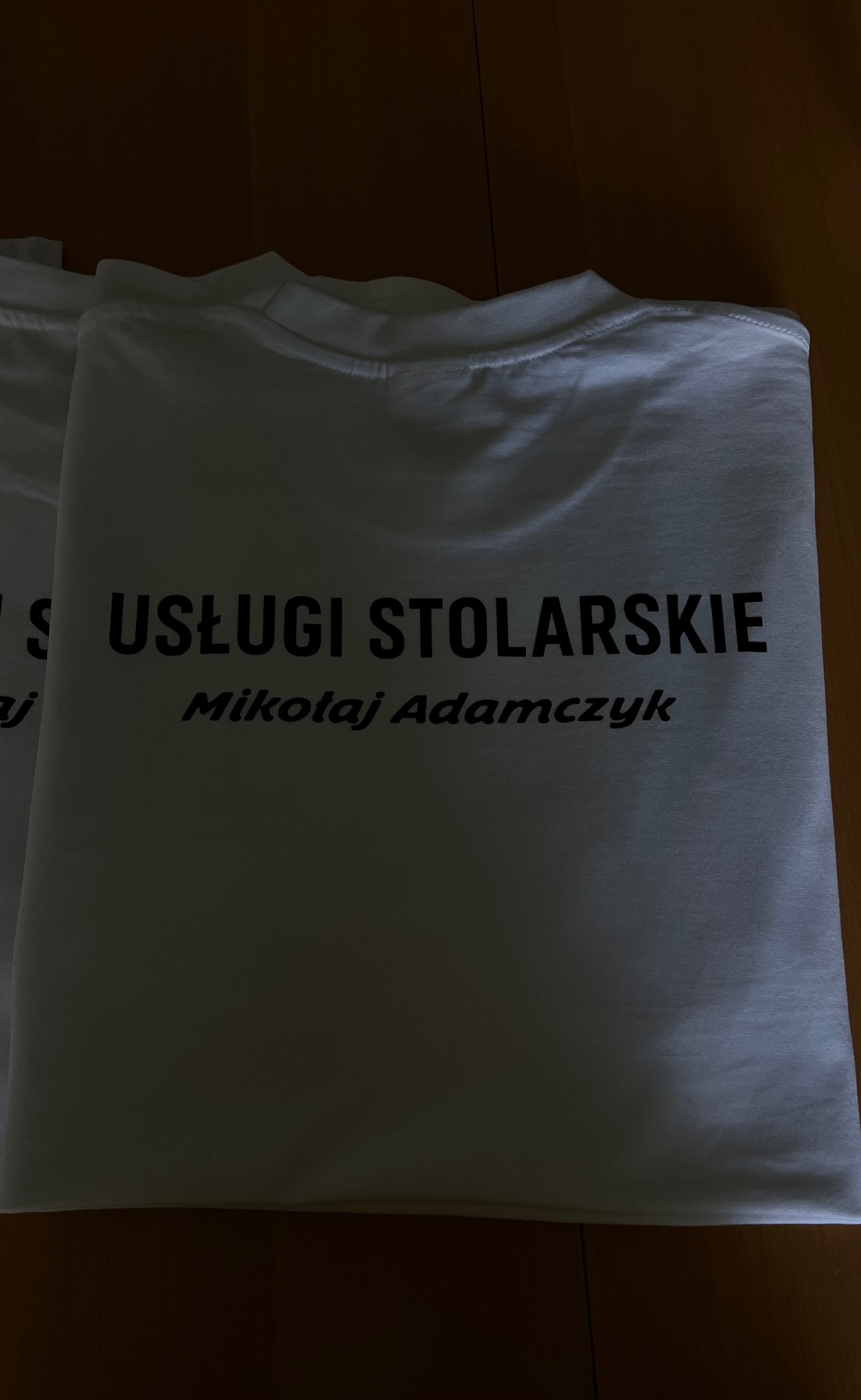 Biała koszulka z czarnym nadrukiem 'Usługi Stolarskie Mikołaj Adamczyk', leżąca na drewnianej powierzchni, widoczna częściowo druga koszulka.