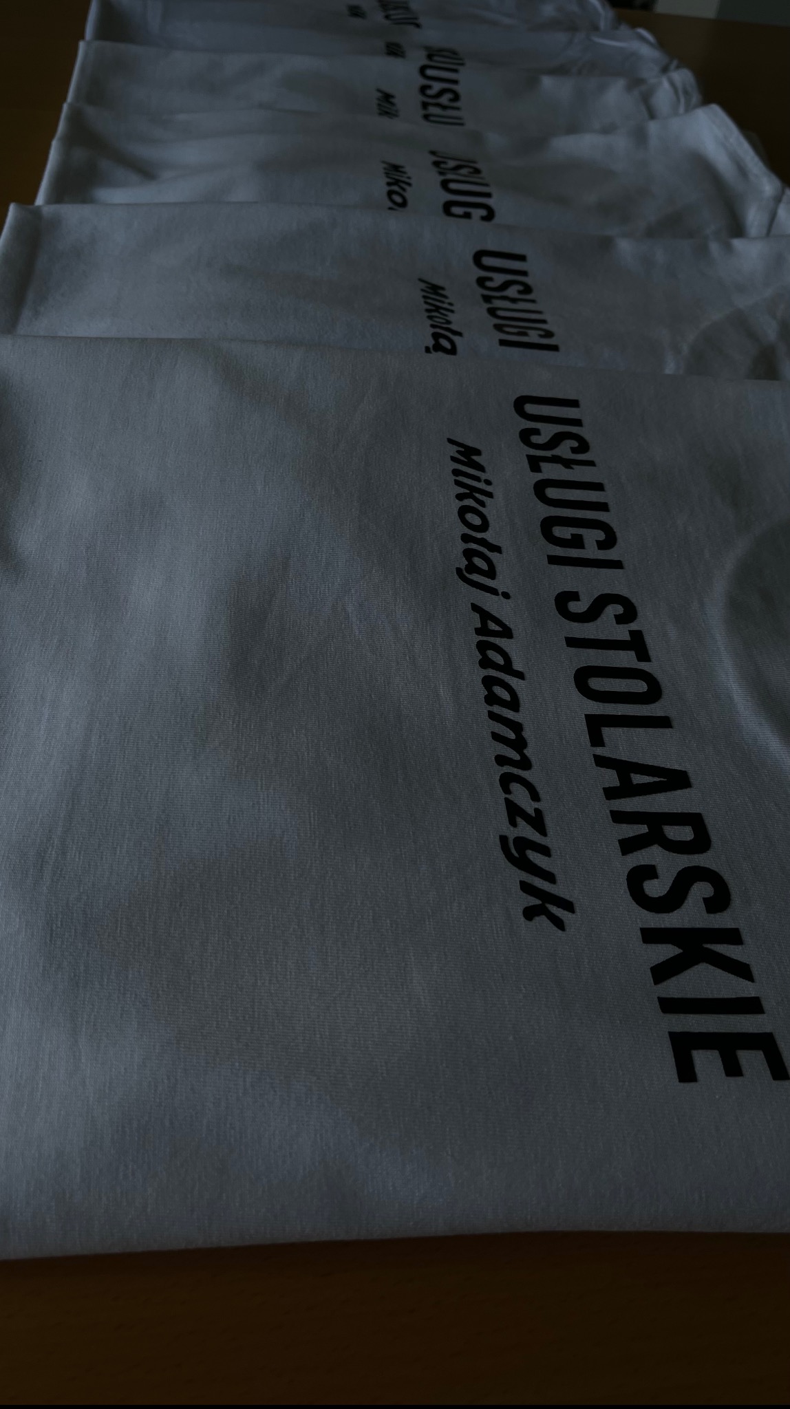 Stos białych koszulek z czarnym nadrukiem 'USŁUGI STOLARSKIE Mikołaj Adamczyk' w perspektywie z góry, ukazujący detale tkaniny i precyzję wykonania nadruku.