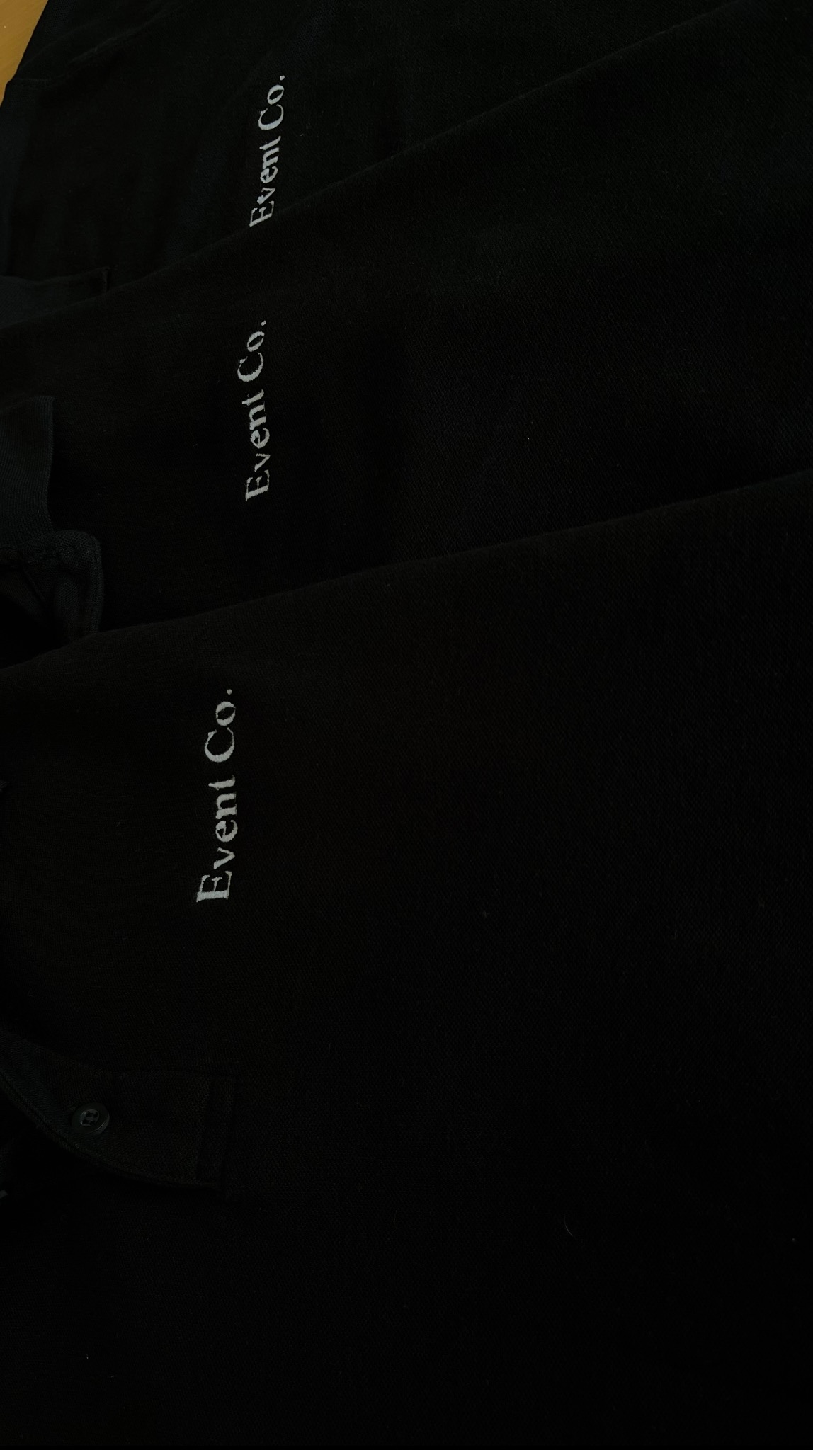 Czarne koszulki polo z białym haftem 'Event Co.', ułożone jedna na drugiej, prezentujące detale wykonania i jakość materiału. Widoczne guziki i fragment kołnierzyka.