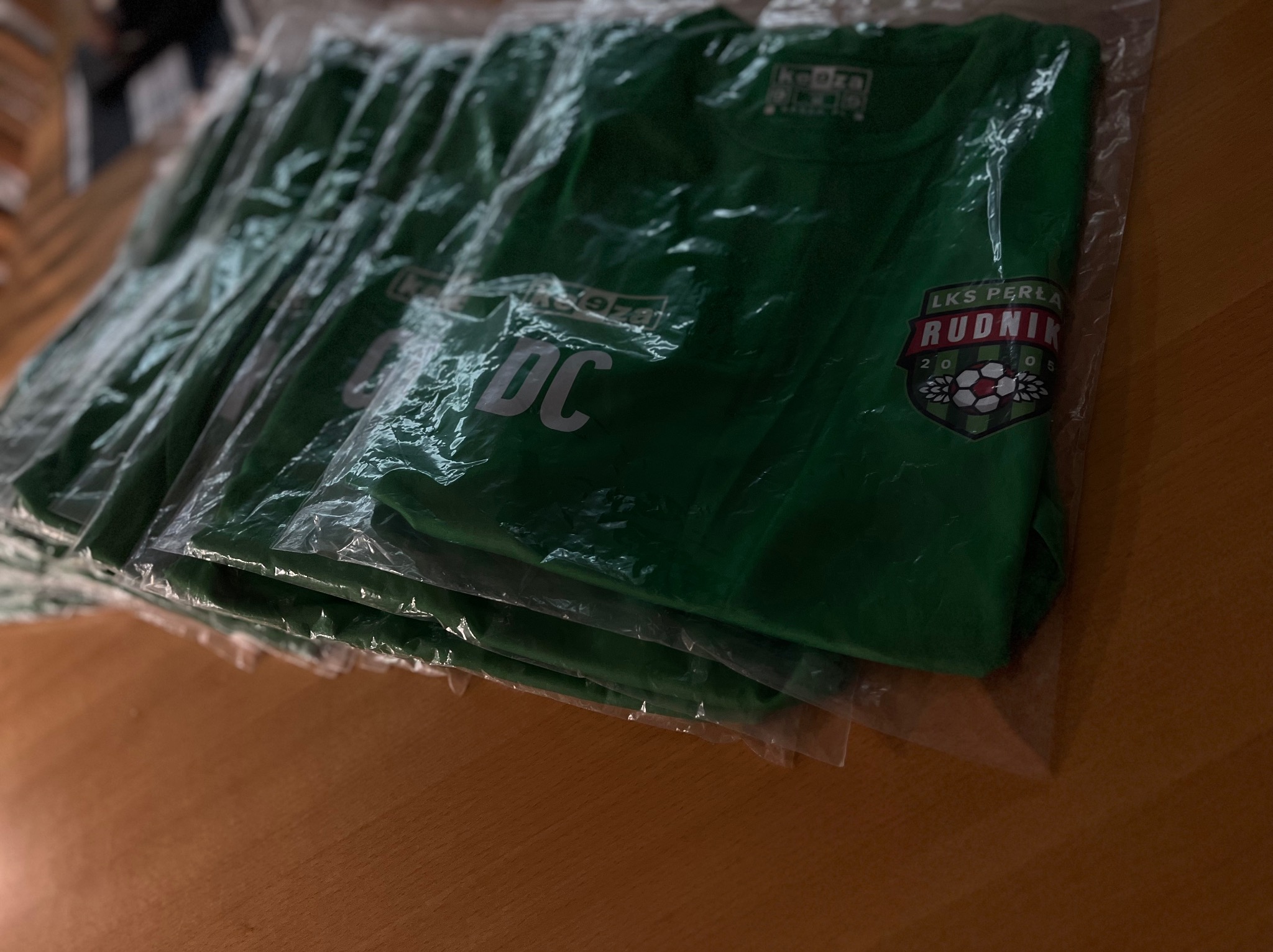 Stos zielonych koszulek w foliowych opakowaniach z nadrukiem logo LKS Perła Rudnik i napisem DC, leżących na drewnianym blacie. Widoczne detale nadruku.