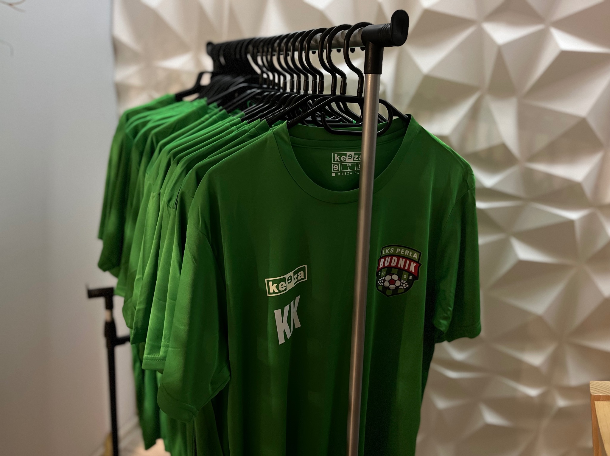 Rząd zielonych koszulek sportowych z nadrukiem logo klubu LKS Perła Rudnik, wiszących na czarnym wieszaku na tle białej, geometrycznej ściany.