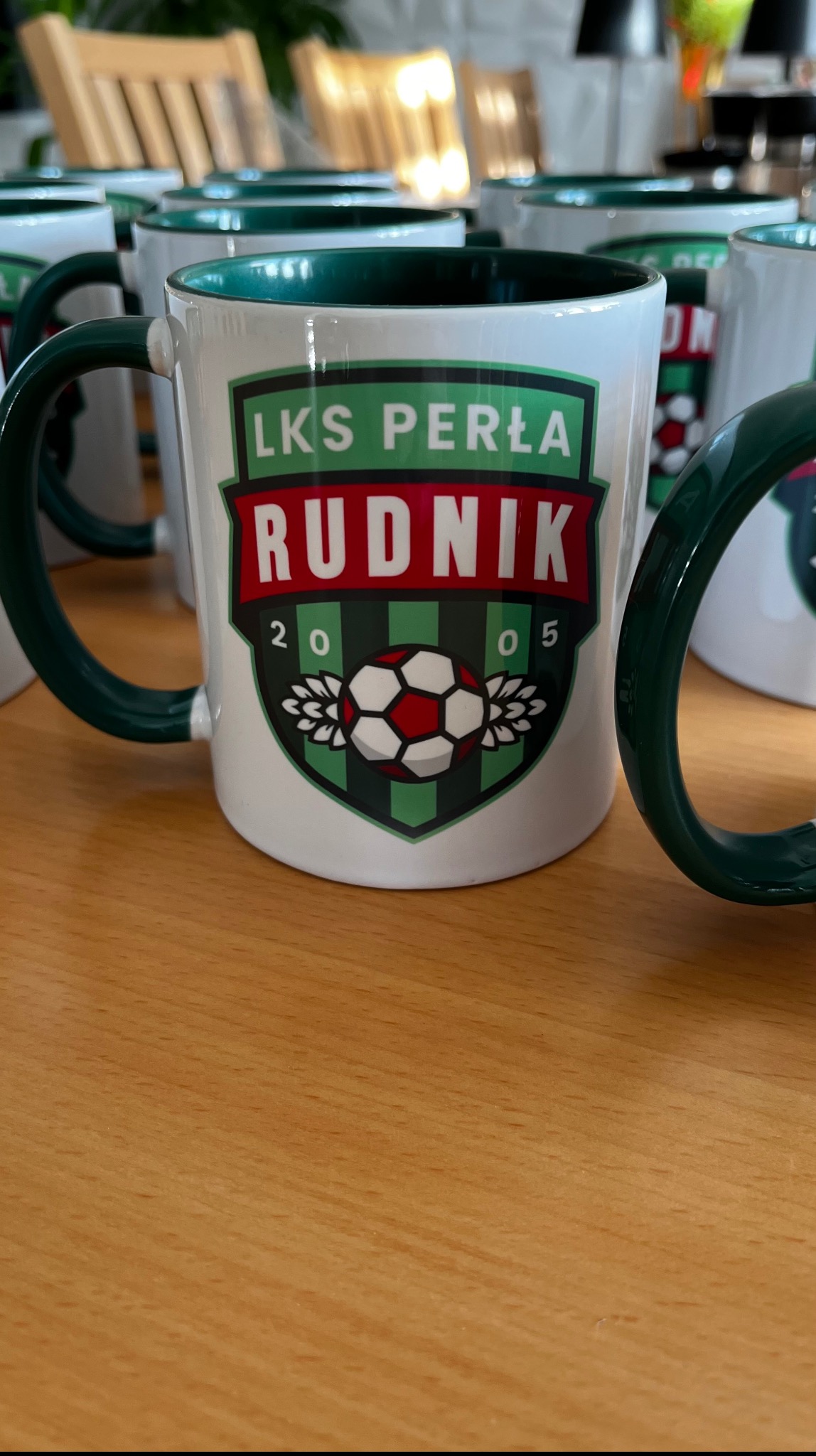 Biały kubek z zielonym wnętrzem i uchwytem, z nadrukiem herbu LKS Perła Rudnik, ustawiony na drewnianym stole, w tle inne kubki.