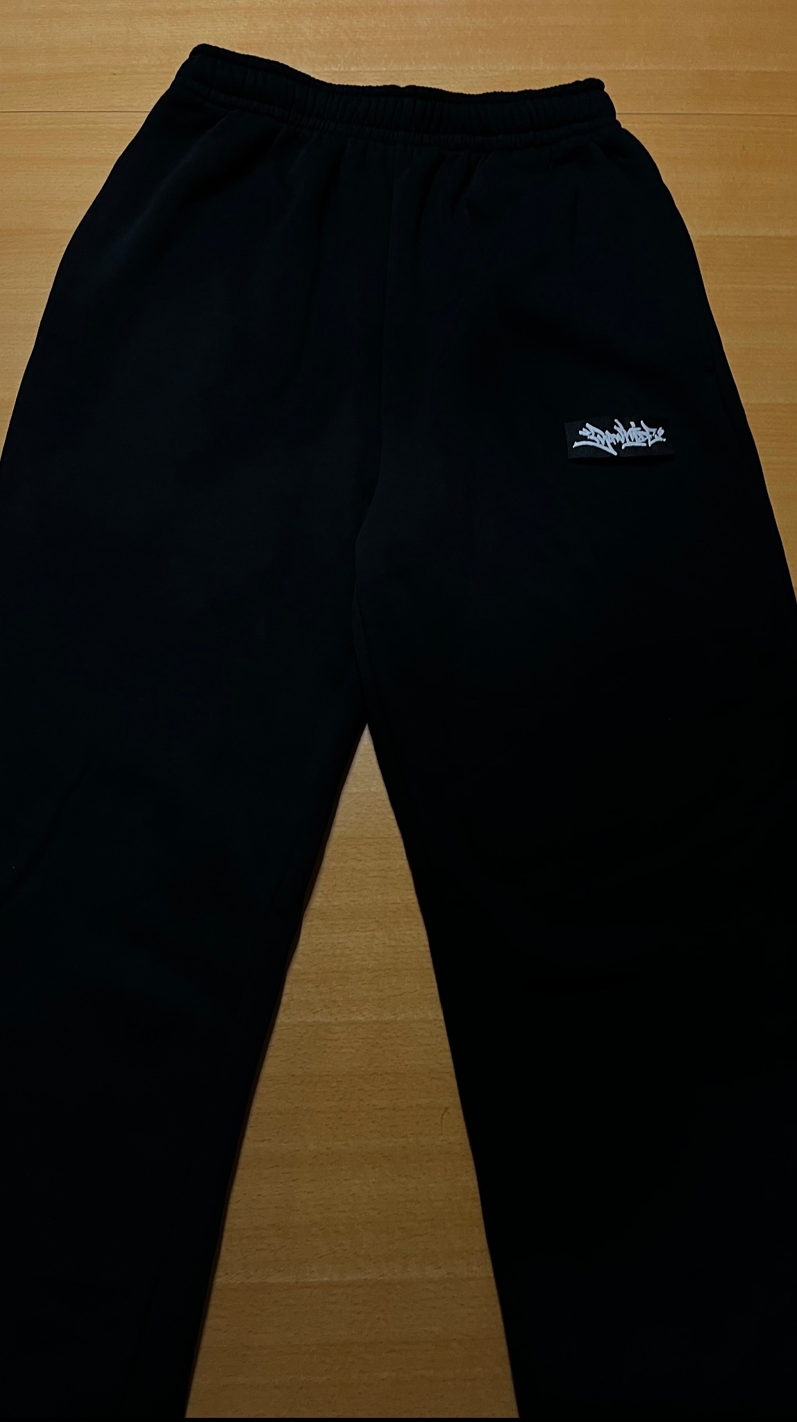 Czarne spodnie dresowe ze sznurkiem w pasie i białym, abstrakcyjnym nadrukiem logo na prawej nogawce, leżące na drewnianej powierzchni. Widok z góry.