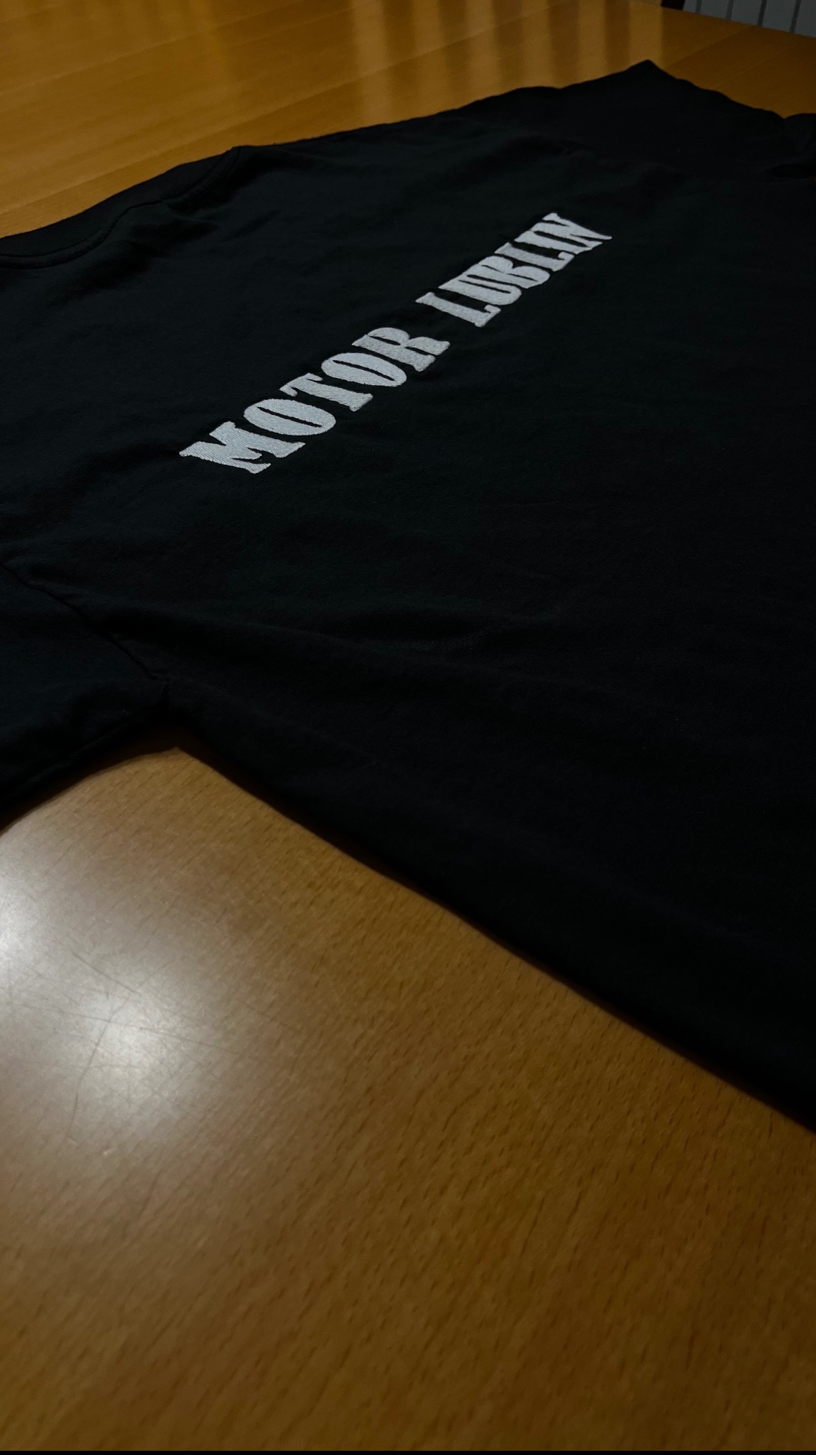 Czarna koszulka z białym nadrukiem 'MOTOR LUBLIN' leżąca na drewnianym stole. Ujęcie z góry, z widocznym fragmentem rękawa i częścią nadruku.