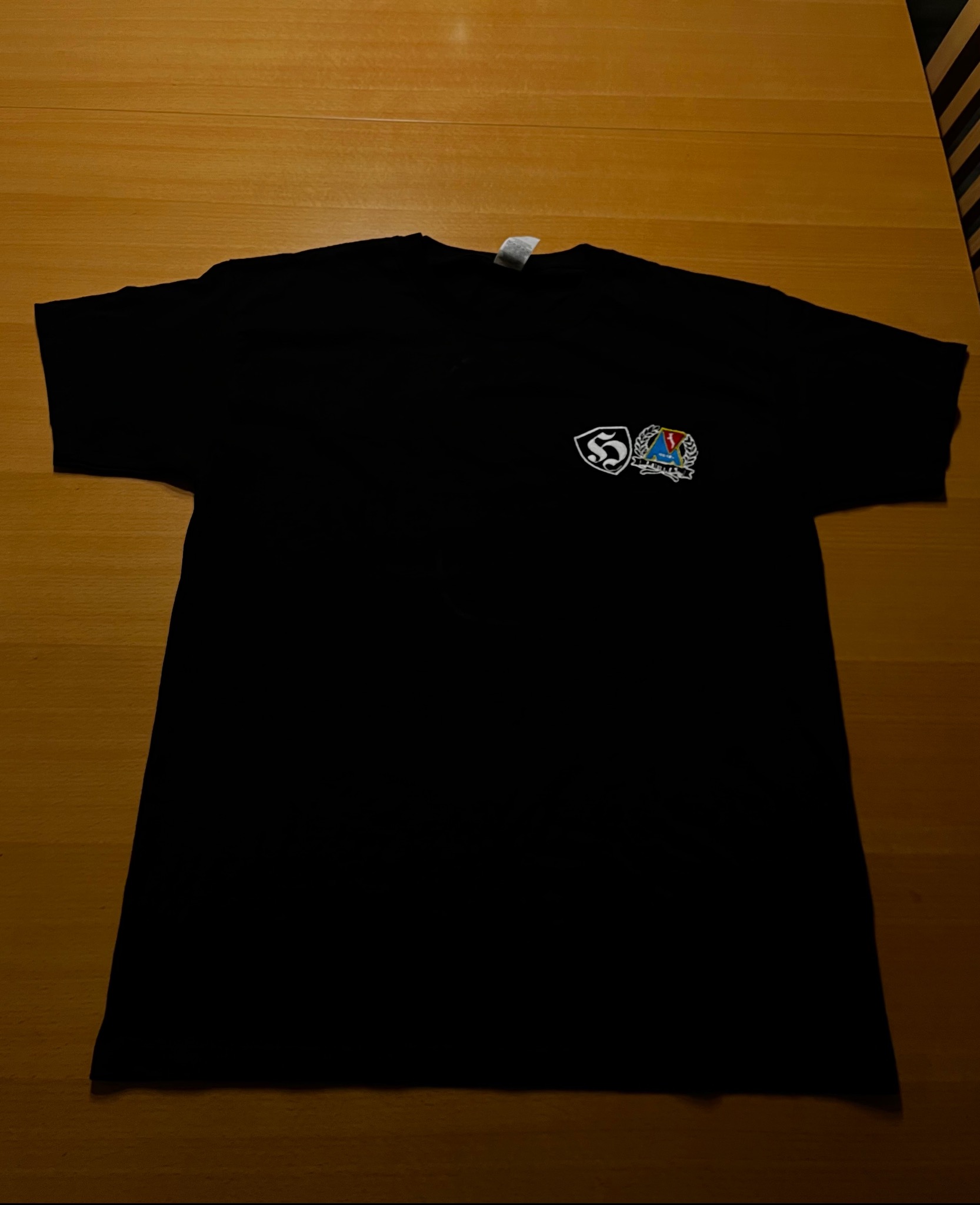 Czarna koszulka z nadrukiem dwóch logotypów na piersi, leżąca na drewnianym stole. Widoczne detale haftu i kolorystyka logo.