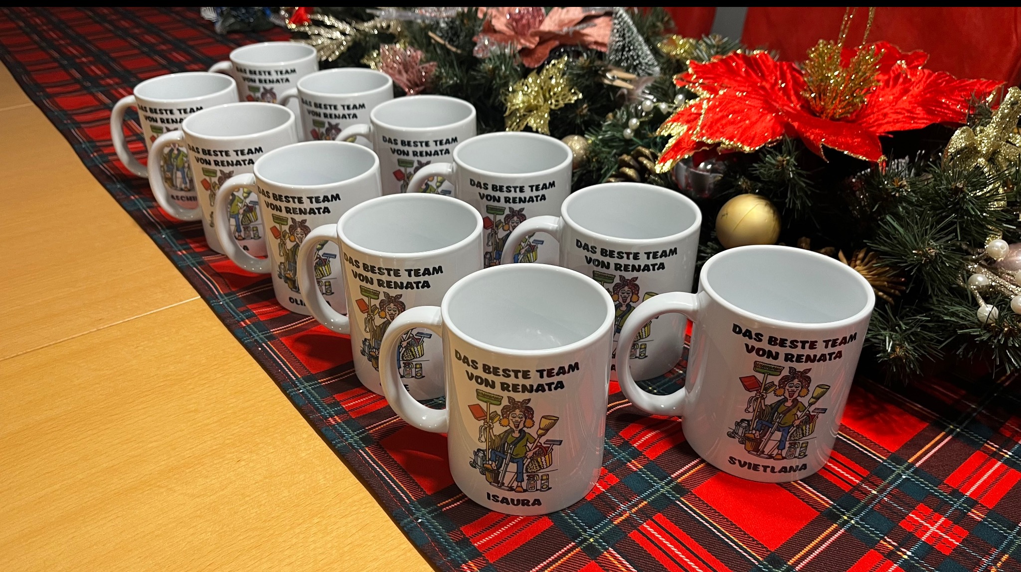 Rząd białych kubków z nadrukiem 'Das Beste Team von Renata' i imionami, ustawionych na czerwono-zielonej kracie, z dekoracjami świątecznymi w tle.
