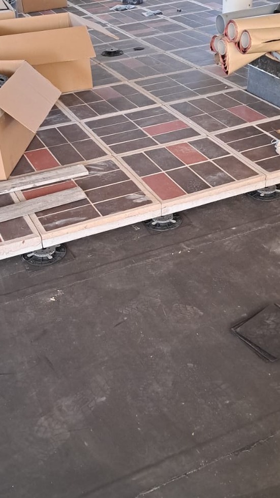 Podniesiona podłoga z kwadratowych płytek w odcieniach brązu i czerwieni, wsparta na regulowanych wspornikach. Widoczne kartony i materiały budowlane w tle.