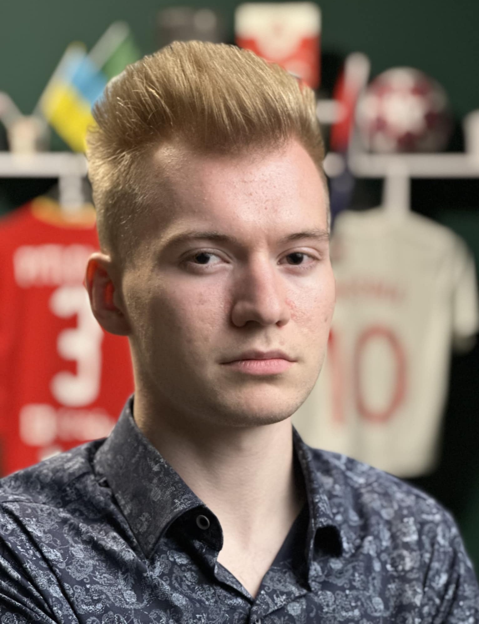 Portret młodego mężczyzny w koszuli z wzorem, z tłem zawierającym koszulki sportowe i flagi, sugerującym zainteresowania sportowe lub tematyczną imprezę.