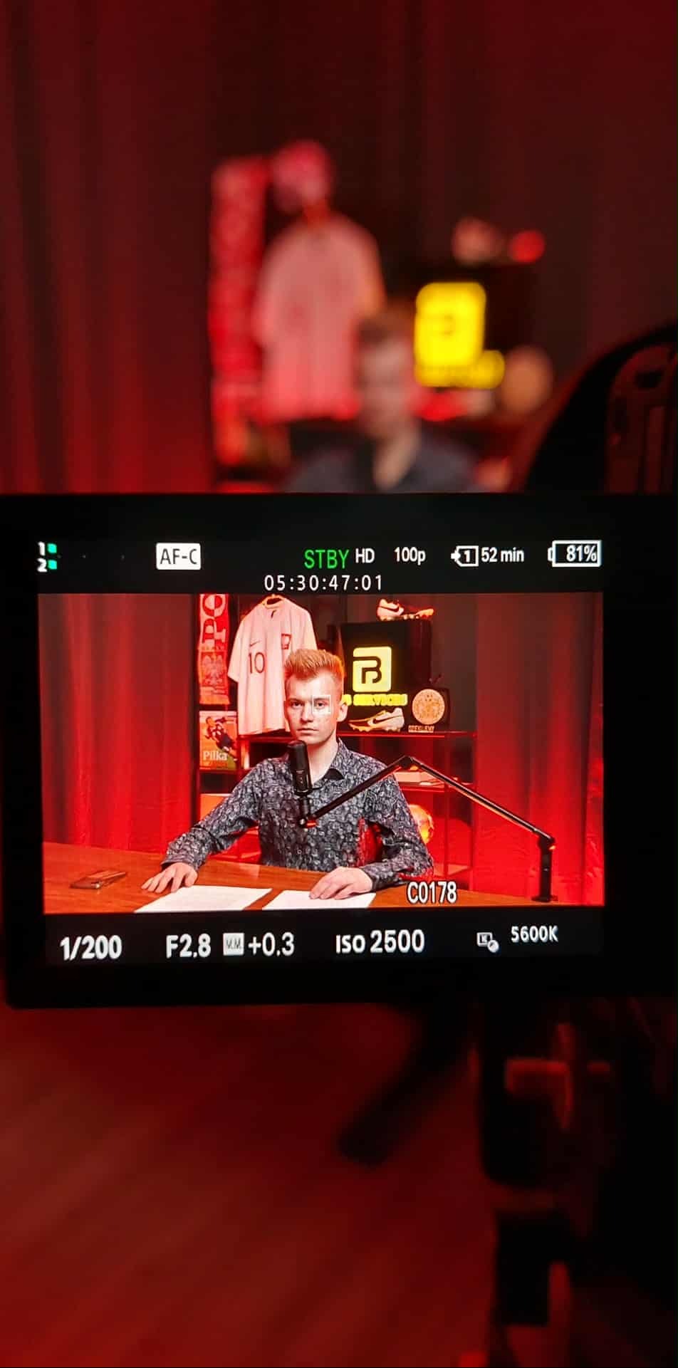 Ekran aparatu z widokiem na młodego mężczyznę nagrywającego w Witanowicach. Widoczne ustawienia ekspozycji i elementy scenografii w tle. Czerwone zasłony.