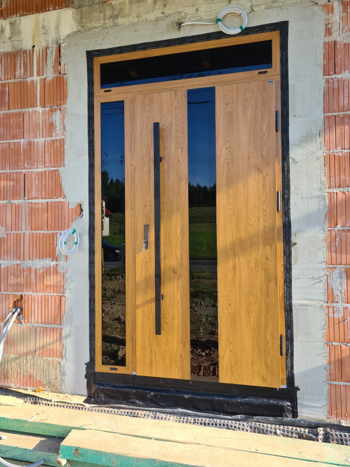 Zewnętrzne drzwi wejściowe z przeszkleniami, w odcieniach drewna, zamontowane w surowym murze. Widoczne detale wykończenia i czarna, długa klamka.