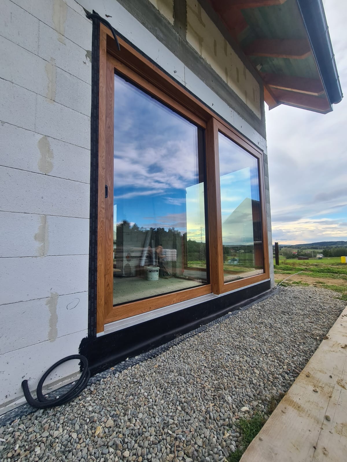 Zamontowane, brązowe drzwi przesuwne z widokiem na niebo i okolicę, w surowym stanie budynku. Widoczne detale montażowe i otoczenie budowy.