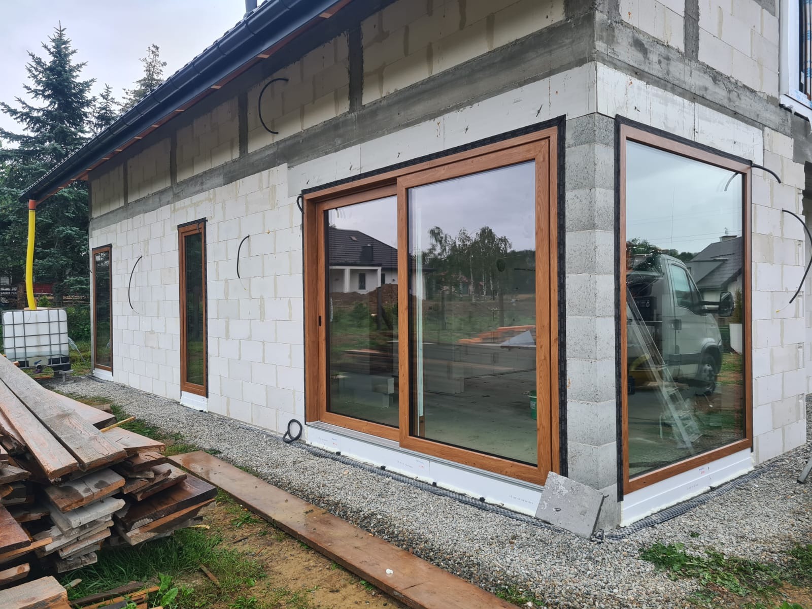 Budynek w trakcie budowy z zamontowanymi oknami i drzwiami przesuwnymi z brązowymi ramami. Widoczne bloczki betonowe i przygotowany teren.