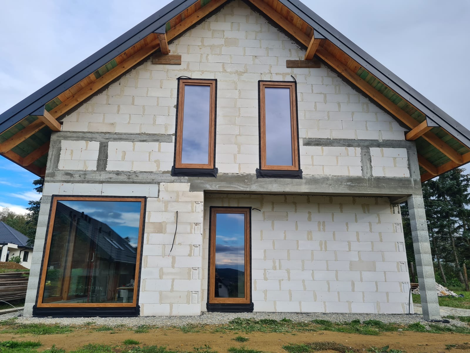 Surowy stan domu z zamontowanymi oknami z drewnianą ramą. Widoczne bloczki, betonowe nadproża i drewniana konstrukcja dachu. W oknach odbija się niebo.