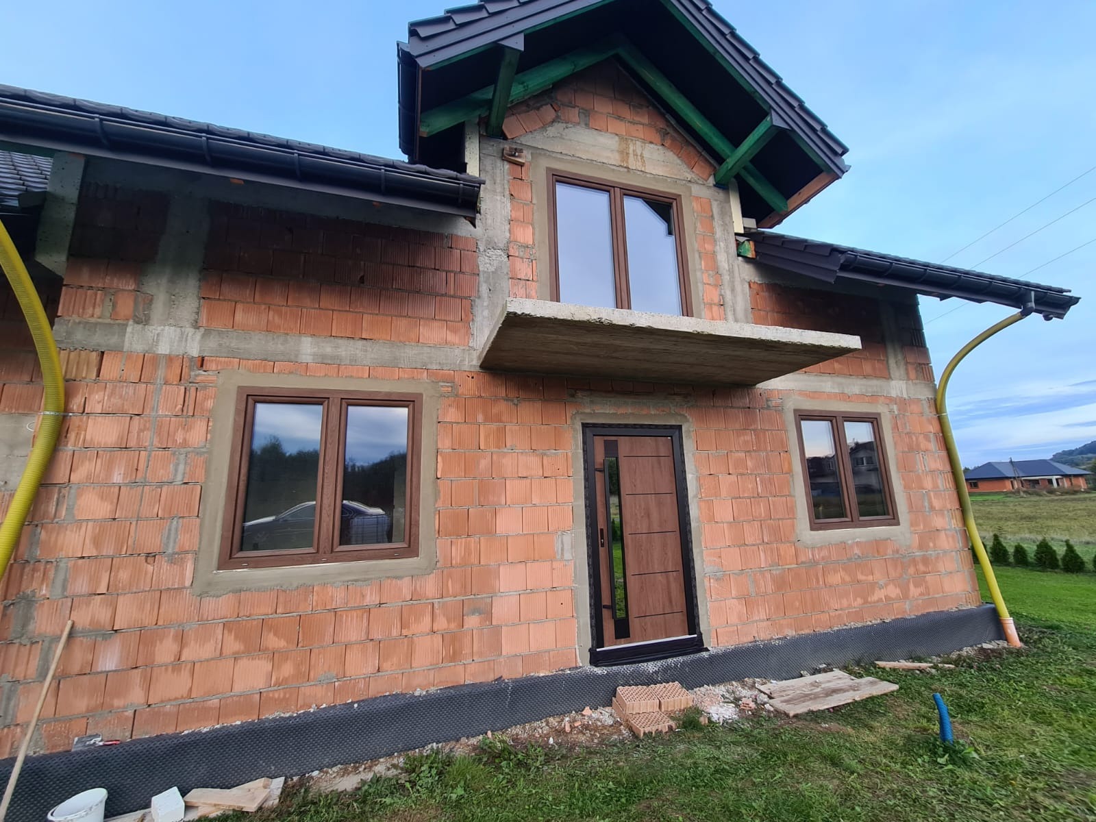 Surowy budynek z brązowymi oknami i drzwiami, widoczne cegły i beton, w tle zielona trawa i inne budynki. Etap budowy z zamontowaną stolarką okienną i drzwiową.