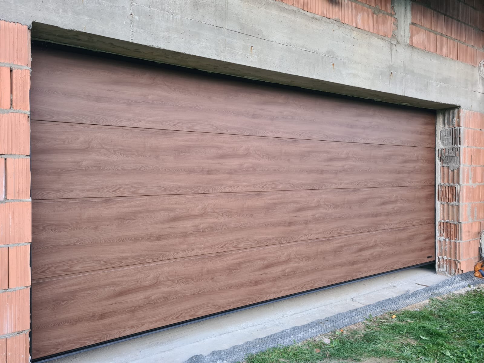 Zamknięta, segmentowa brama garażowa w kolorze drewna, zamontowana w surowym otworze budynku z cegły i betonu. Widoczne detale konstrukcyjne.