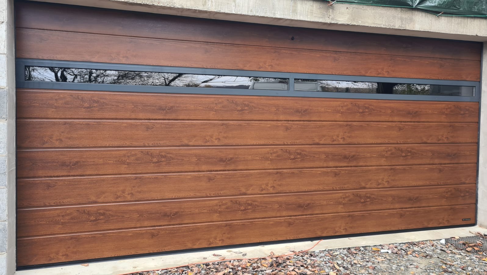 Segmentowa brama garażowa w odcieniach brązu z poziomymi panelami i wąskim oknem, odbijającym drzewa. Widoczne betonowe elementy konstrukcyjne.