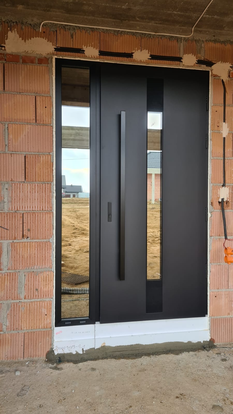Czarne drzwi zewnętrzne z przeszkleniami, zamontowane w surowym murze z czerwonej cegły. Długi, pionowy uchwyt. Widoczne przewody elektryczne.