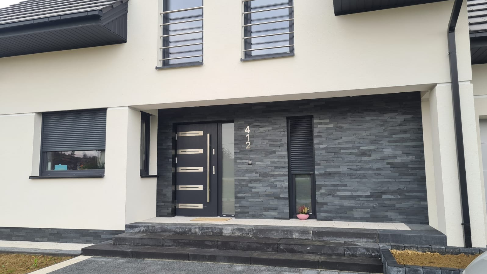 Elewacja domu z szarą stolarką: drzwi z numerem 412, roleta na oknie, okna z metalowymi kratami. Nowoczesny design, grafitowa cegła elewacyjna.