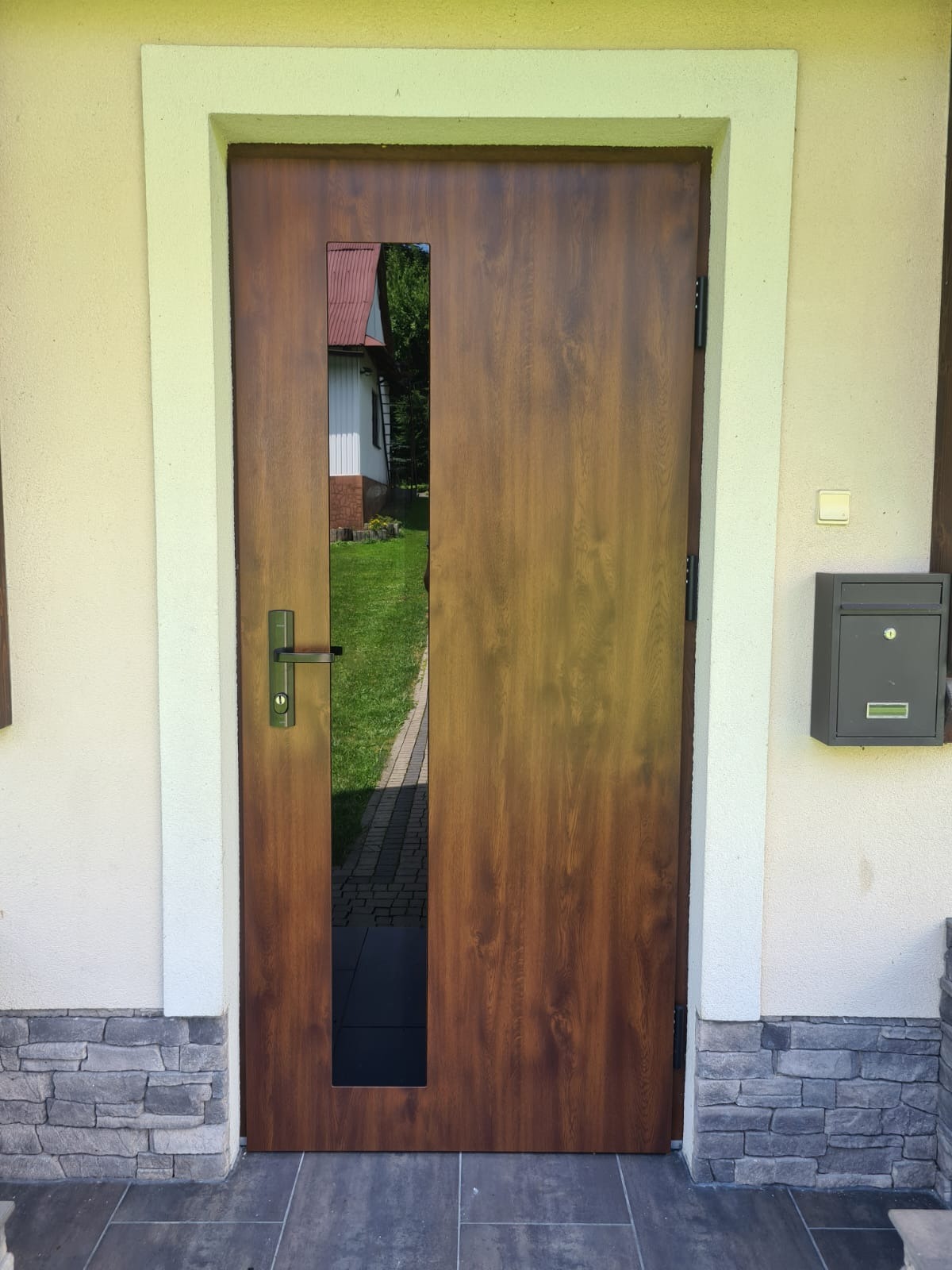 Brązowe drzwi wejściowe z wąskim, pionowym przeszkleniem, zamontowane w jasnej elewacji z kamienną podmurówką. Widoczna skrzynka na listy i fragment ogrodu w odbiciu.