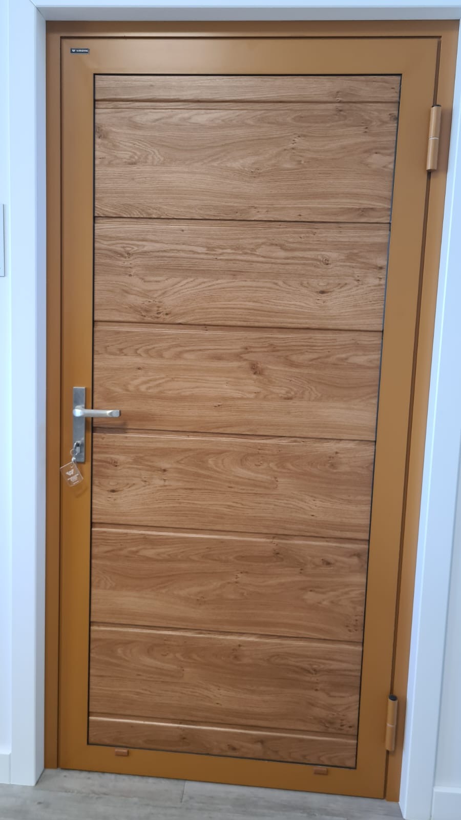 Zewnętrzne drzwi z drewnianym panelem i złotą ramą, widoczne z bliska, z kluczem w zamku. Minimalistyczny design, ciepłe oświetlenie.