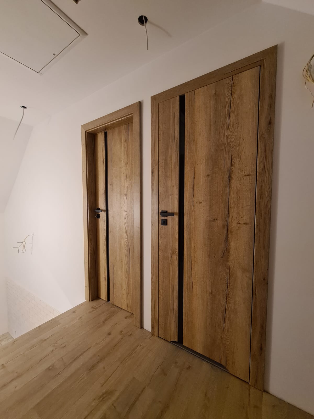 Dwa drewnopodobne skrzydła drzwiowe z czarnymi akcentami i klamkami, zamontowane w białej ścianie, widoczne na tle podłogi z paneli.