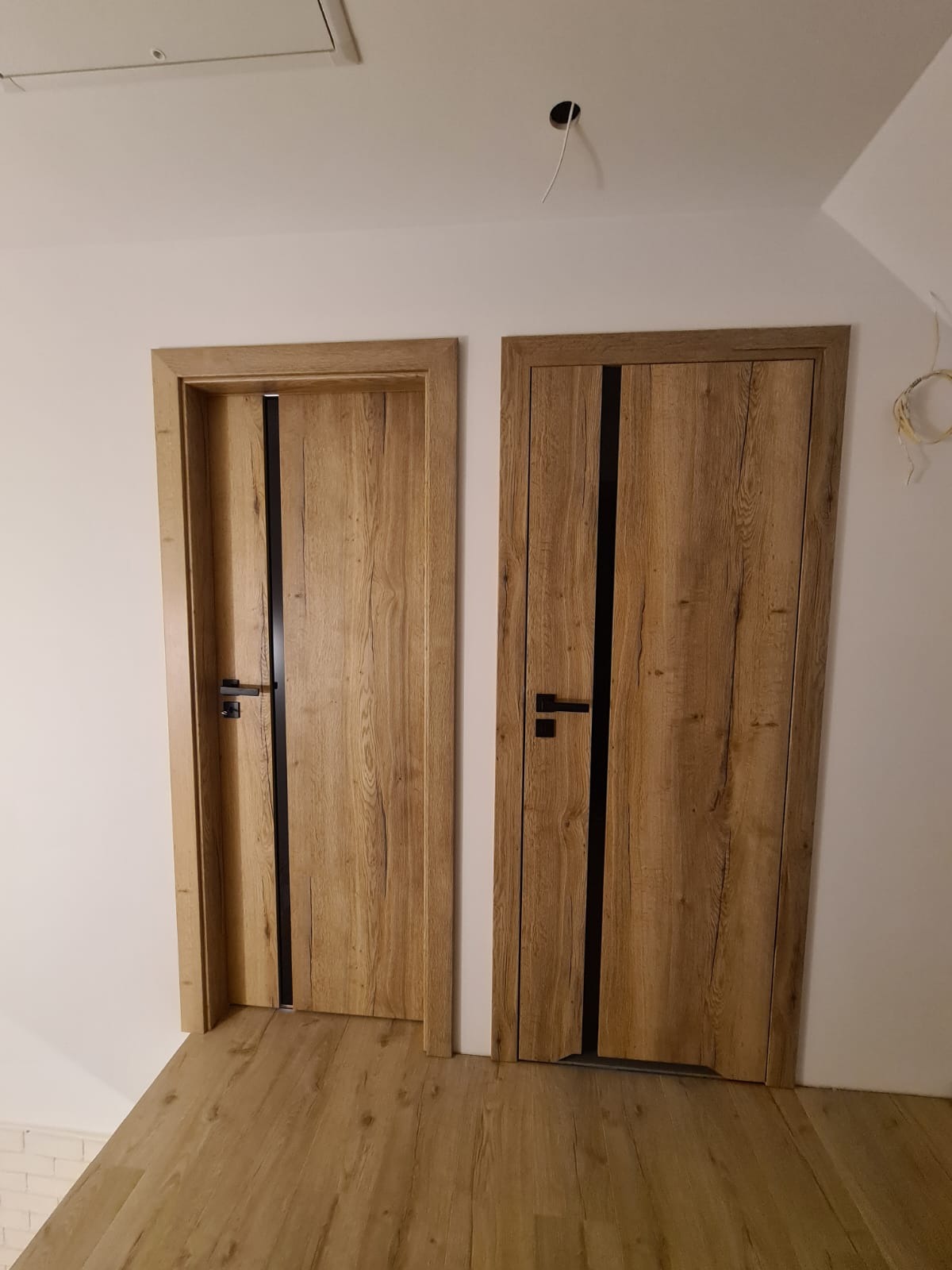 Dwa drewniane drzwi wewnętrzne z czarnymi akcentami, zamontowane w białej ścianie. Podłoga z paneli imitujących drewno. Widoczne niedokończone prace elektryczne.