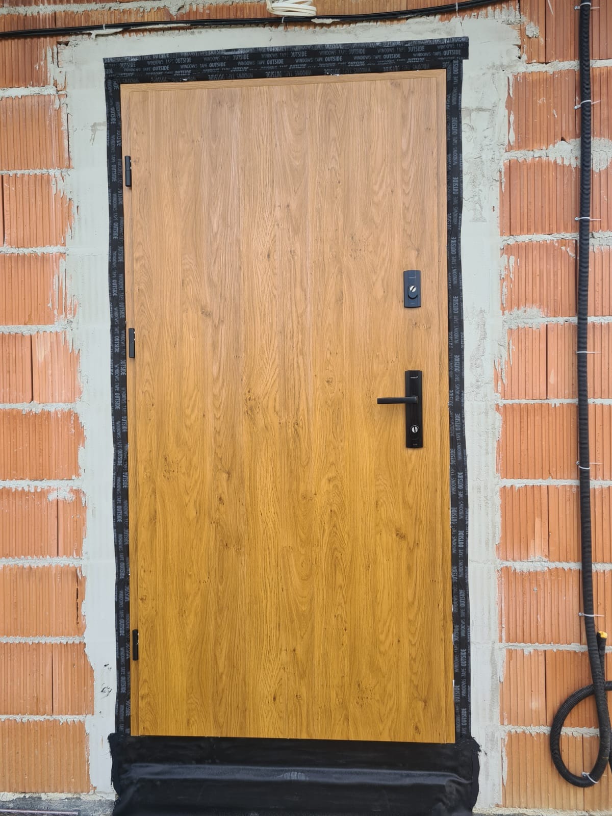 Montaż drzwi zewnętrznych w surowym stanie, widoczne cegły i taśma uszczelniająca. Drzwi w odcieniach brązu z czarną klamką i zamkiem.