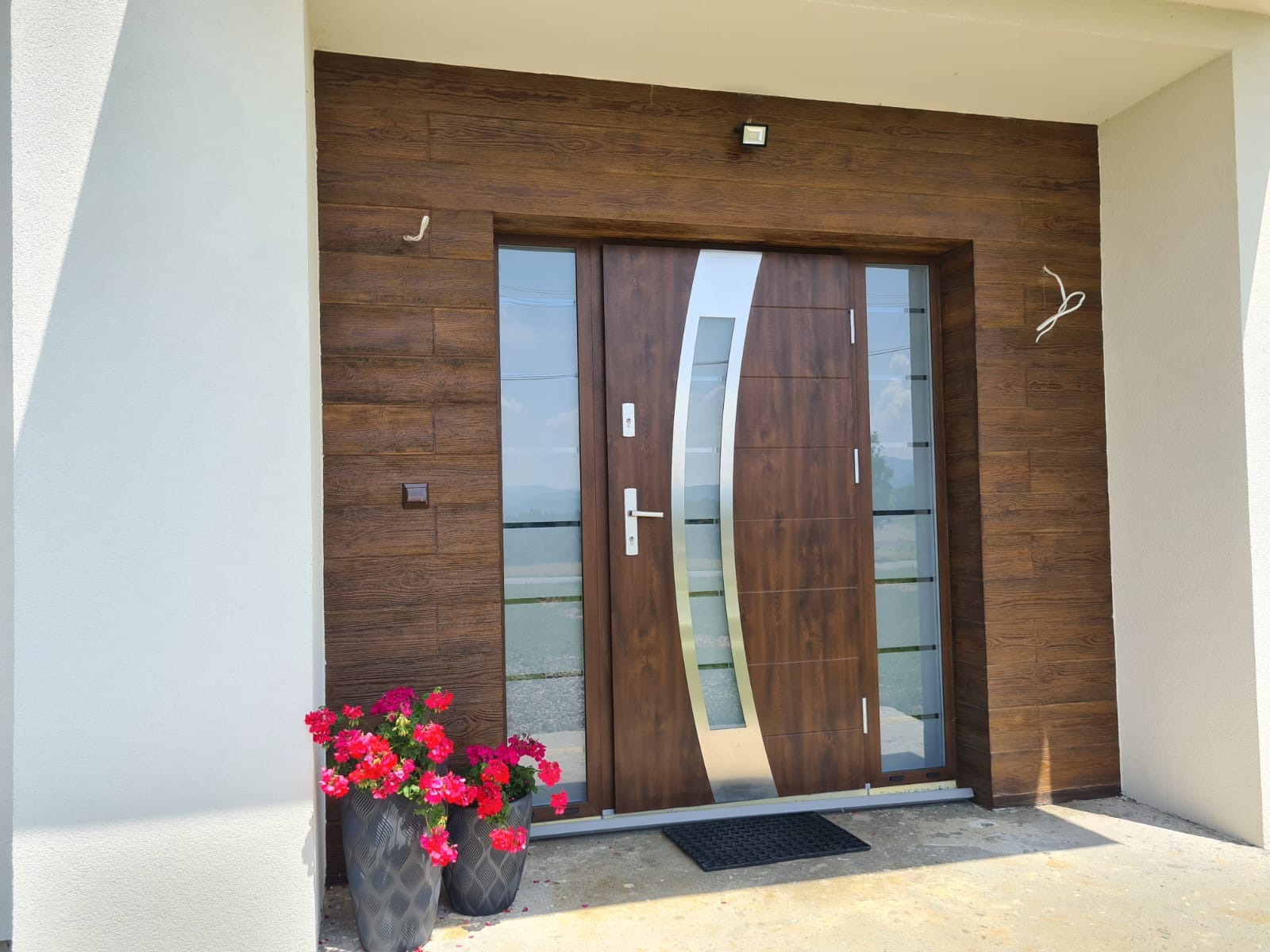 Nowoczesne drzwi zewnętrzne z przeszkleniami i elementami stali, wkomponowane w elewację z drewnopodobną okładziną. Dwa kwiatowe donice dodają koloru.