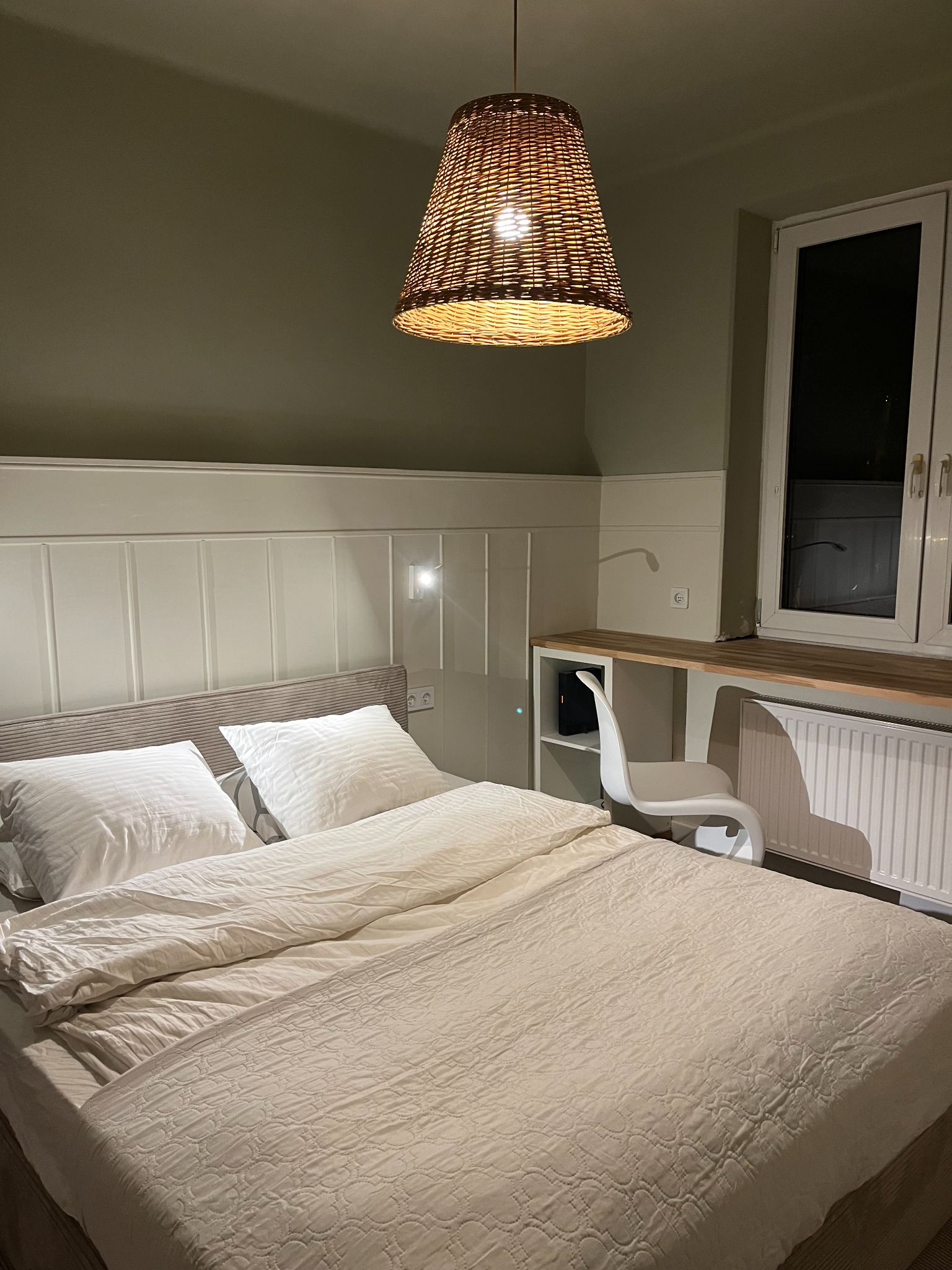 Jasna sypialnia z łóżkiem, biurkiem pod oknem i plecioną lampą. Ściany w odcieniach beżu i zieleni. Minimalistyczny design z elementami naturalnego drewna.