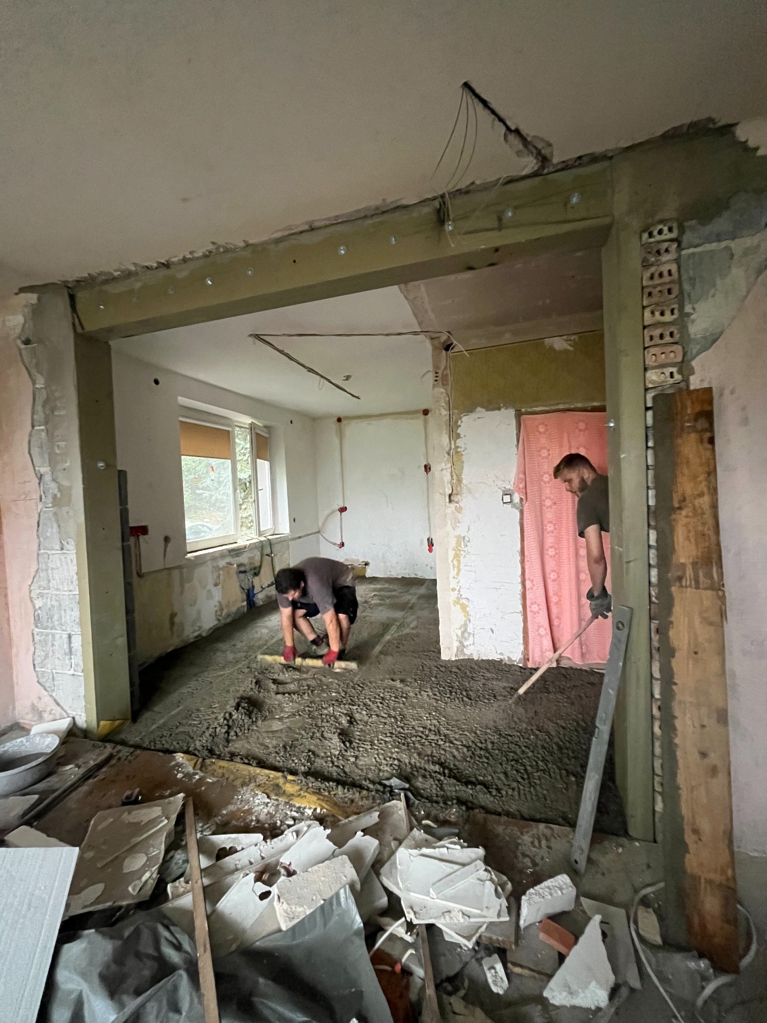 Wyrównywanie wylewki w remontowanym pokoju. Dwóch mężczyzn rozprowadza betonową masę, widoczne surowe ściany i gruz. Etap prac przygotowawczych do dalszych prac wykończeniowych.