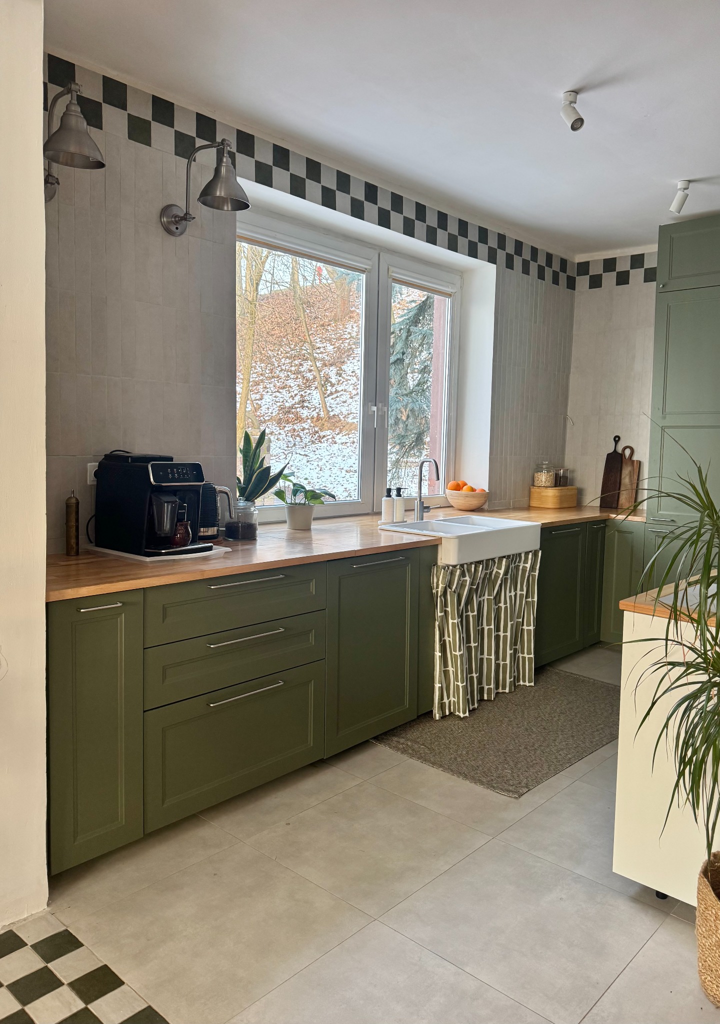 Kuchnia w odcieniach zieleni z drewnianym blatem, białym zlewem i wzorzystą zasłonką. Na blacie ekspres do kawy i dekoracje roślinne. Widoczne płytki w szachownicę na podłodze.