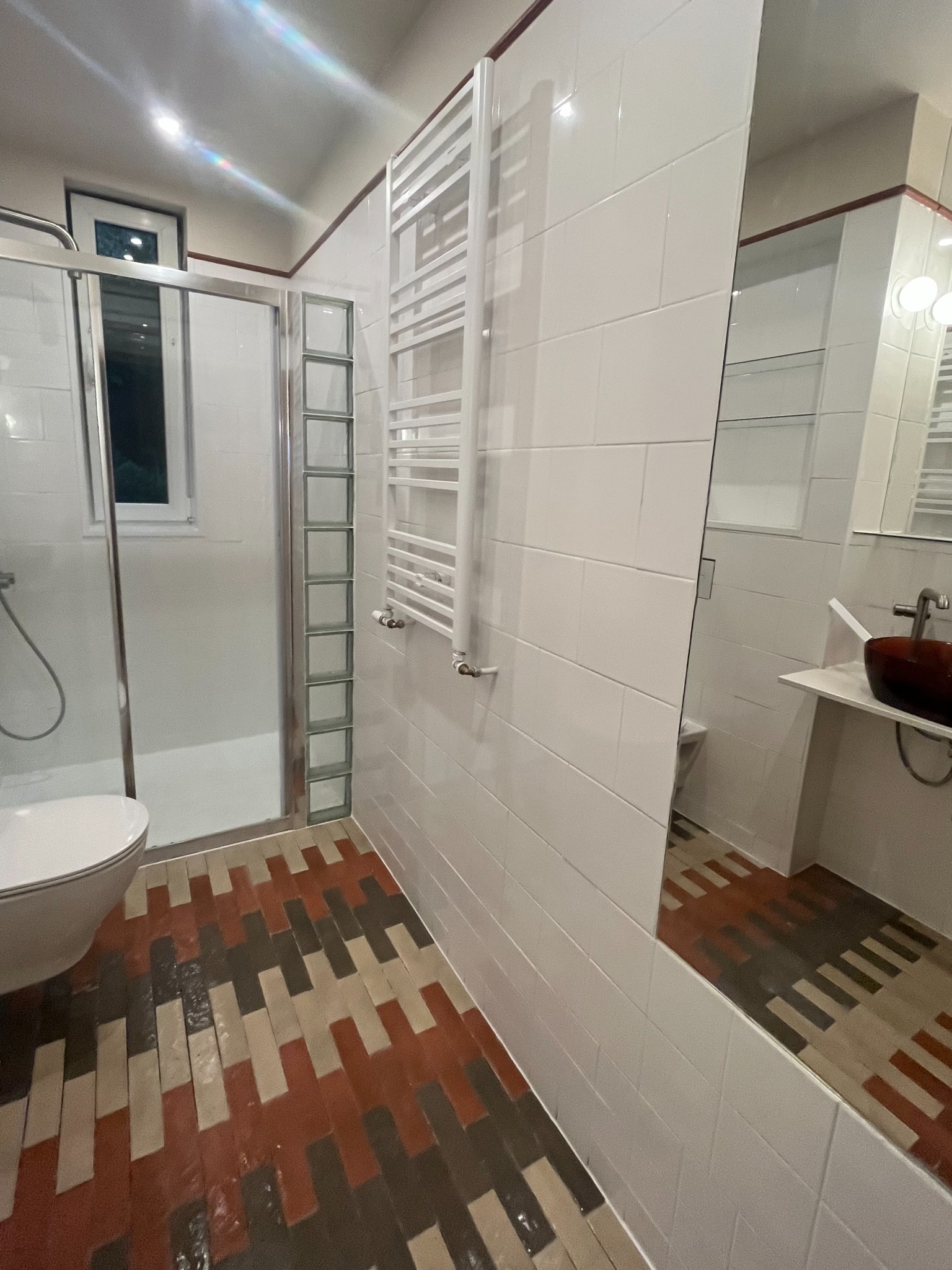 Nowoczesna łazienka z kabiną prysznicową, grzejnikiem drabinkowym i lustrem. Uwagę przyciągają kolorowe płytki podłogowe ułożone we wzór cegiełki.