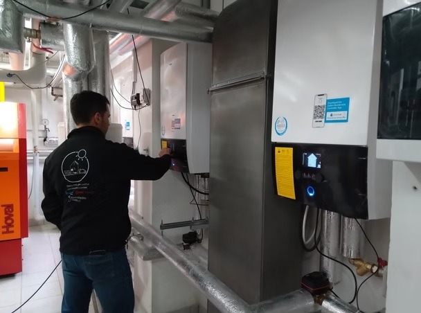 Instalator reguluje nowoczesną pompę ciepła w kotłowni. Widoczne rury, inne urządzenia i logo firmy na ubraniu instalatora.