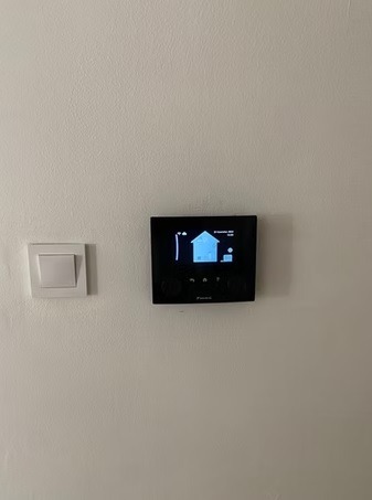 Czarny panel sterowania klimatyzacją Daikin zamontowany na białej ścianie, obok włącznik światła. Na ekranie ikona domu i inne symbole.