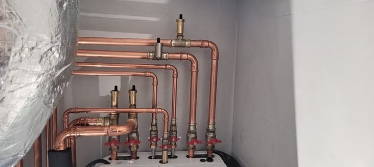 Miedziane rury instalacji grzewczej z zaworami, widoczne elementy kotła i izolacja termiczna. Instalacja w pomieszczeniu o jasnych ścianach.