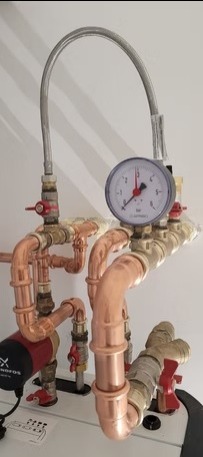 Instalacja hydrauliczna z miedzianymi rurami, manometrem i zaworami, zamontowana na białej ścianie. Widoczna pompa obiegowa. Ujęcie z bliska.