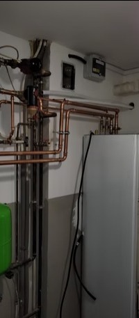 Kompleksowa instalacja grzewcza z miedzianymi rurami, zbiornikiem i licznikiem na ścianie. Widoczne elementy automatyki i sterowania. Realizacja w kotłowni.