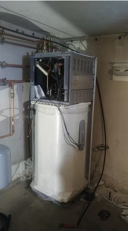 Rozłożona pompa ciepła w trakcie montażu, widoczne rury miedziane, zbiornik i elementy wewnętrzne. Prace instalacyjne w pomieszczeniu technicznym.