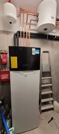 Nowoczesna, biała pompa ciepła w kotłowni z miedzianymi rurami i zbiornikami. Widoczna drabina i elementy instalacji. Minimalistyczny design, szare ściany.