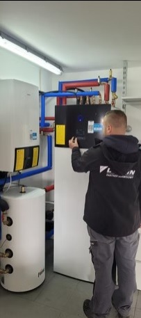 Instalacja pompy ciepła Daikin: instalator w kurtce firmowej reguluje panel sterowania. Widoczne rury w izolacji, zbiornik buforowy i oświetlenie LED.
