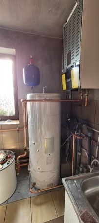 Biały zbiornik buforowy z miedzianymi rurami w pomieszczeniu z oknem i zlewem. Widoczny również wiszący na ścianie element instalacji grzewczej.