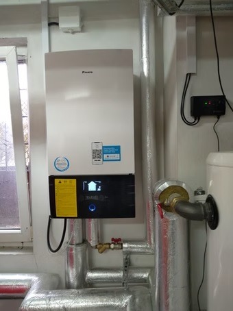 Biała pompa ciepła Daikin z ekranem dotykowym, zamontowana na ścianie w pomieszczeniu technicznym, widoczne rury i elementy instalacji.