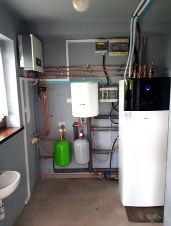 Kompletna instalacja pompy ciepła w pomieszczeniu technicznym: biała jednostka, zbiorniki wyrównawcze (zielony i szary), miedziane rury, licznik energii, biała umywalka.