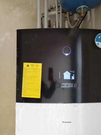 Nowoczesna pompa ciepła Daikin z wyświetlaczem i żółtą naklejką ostrzegawczą, widoczne rury instalacji grzewczej w tle. Kompaktowe rozwiązanie dla domu.